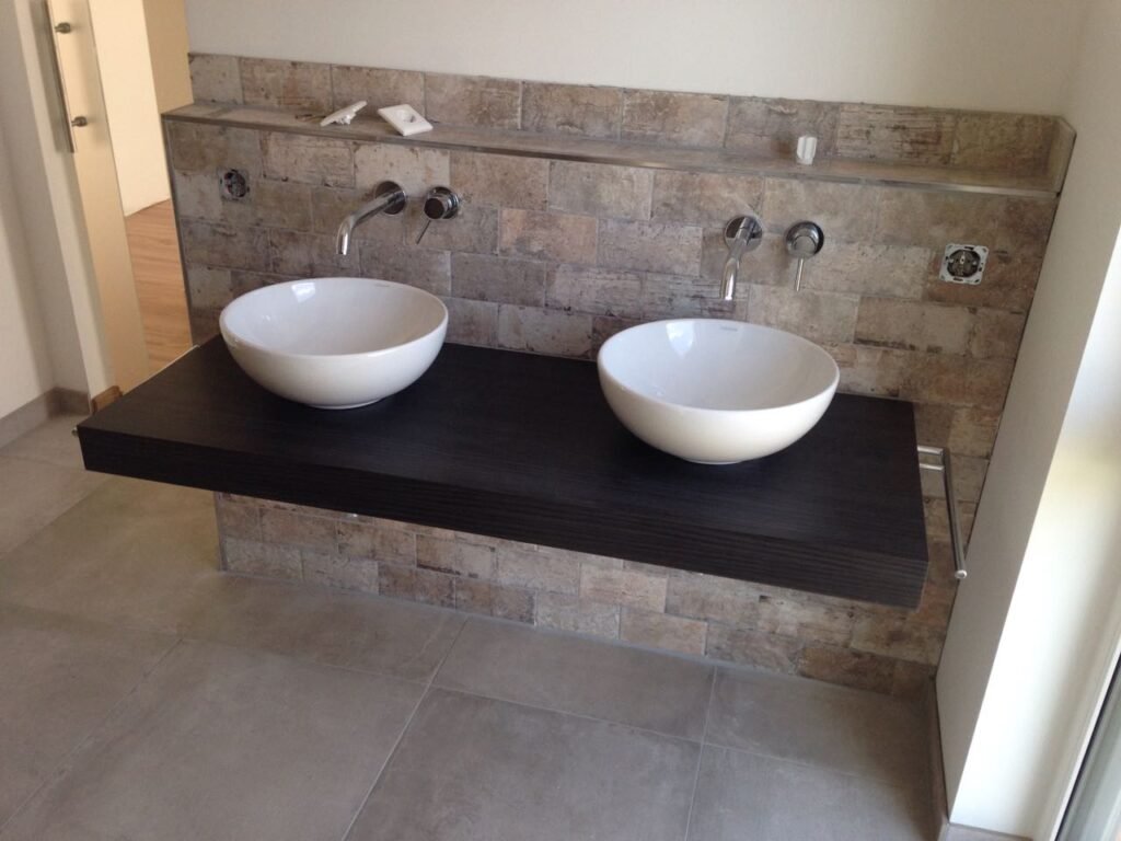 Zwei Aufsatzbecken auf schwebendem Holzregal vor rustikaler Fliesenrückwand und wandmontierten Armaturen, Badsanierung, Fliesenarbeiten