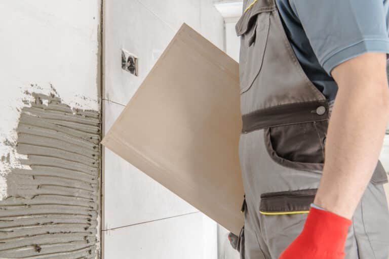 Handwerker hält Wandfliese, trägt Fliesenkleber auf und setzt Fliesen ein, Fliesenarbeiten bei Badsanierung